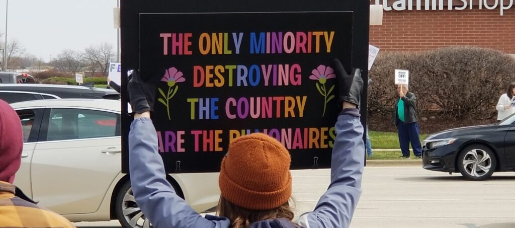 Prostester photographed from behind, holding a poster with multi colored lettering that says:
The Only Minority Destroying our Country are the Billionaires.