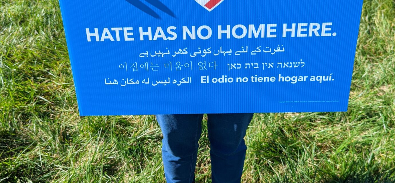 A blue poster with a heart colored like the US Flag. Motto on the poster says Hate has no Home Here.